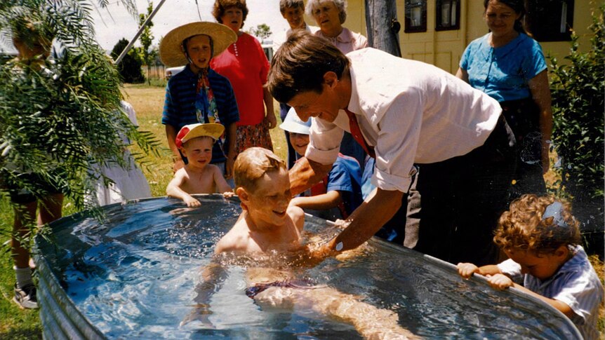A man in a white shirt stands over a boy in an outdoor bath