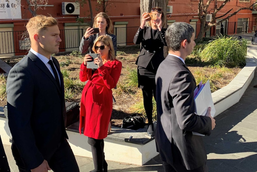 A young man and two older men, all wearing suits, walk past three women taking photos with their smartphones.