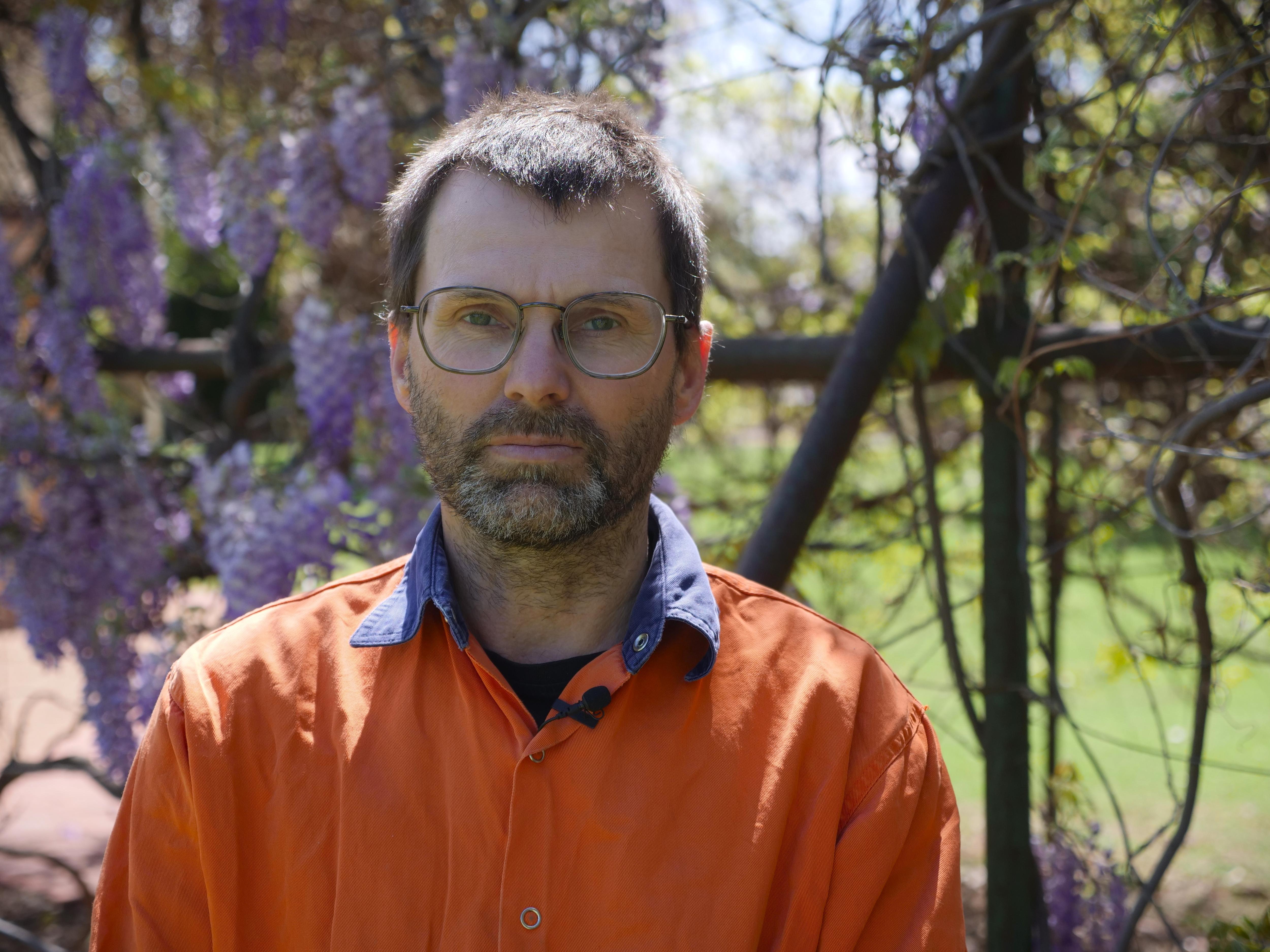 A middle-aged, bearded man with dark hair stands near a wisteria-covered trellis.