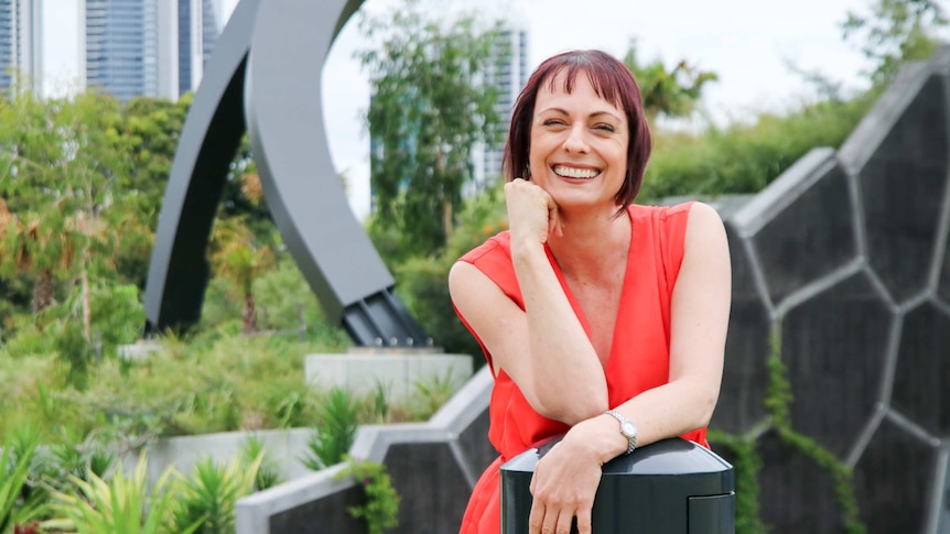 A smiling woman with short red hair, wearing a red dress, standing in a city public garden.