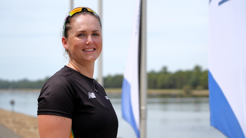 A female Paralympian stands and smiles at the camera, with a lake and flags in the background.