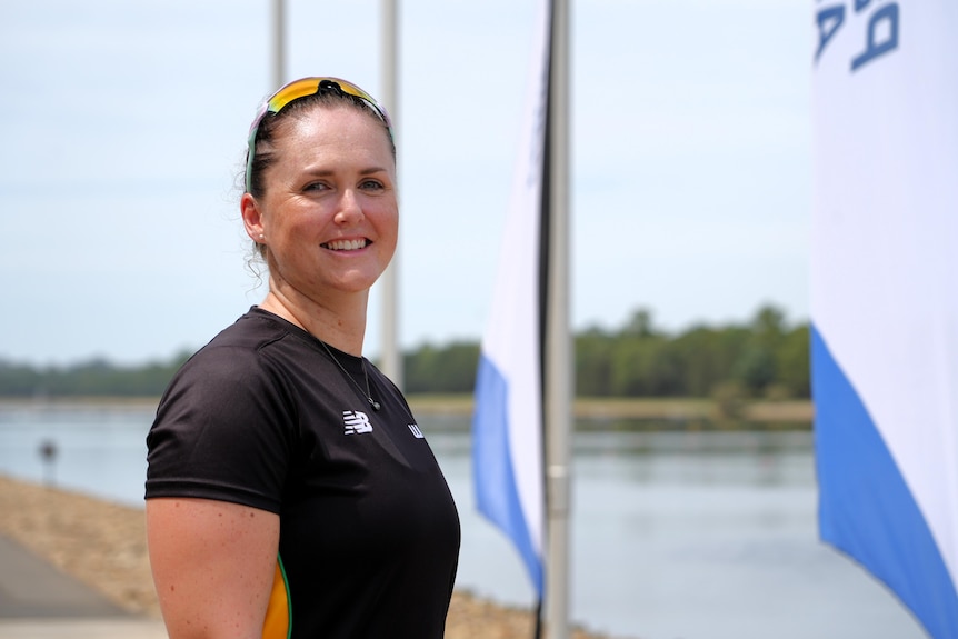 A female Paralympian stands and smiles at the camera, with a lake and flags in the background.