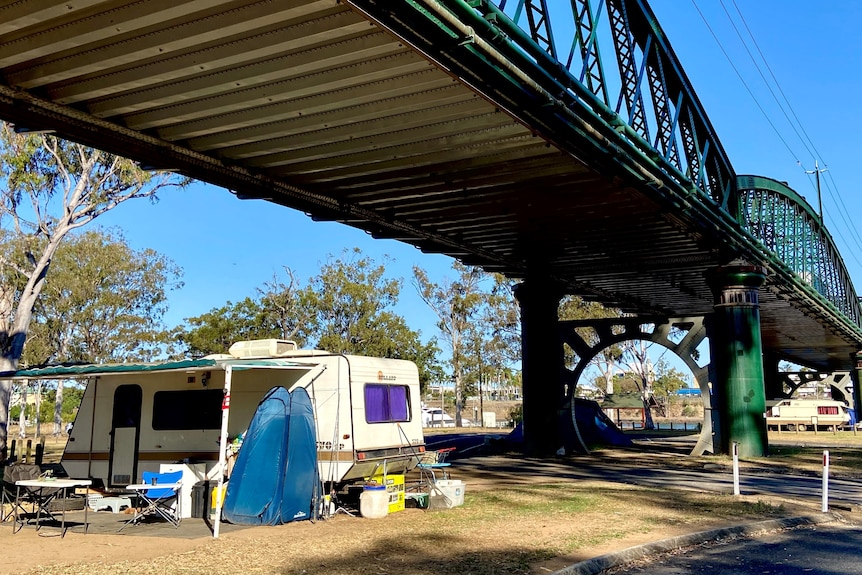 A campsite under a large traffic bridge. Parkland in the background, with other campsites at back.