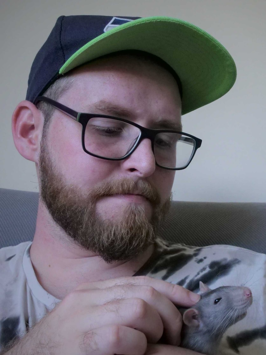 Man wearing cap sitting on couch patting a pale grey pet rat on the head