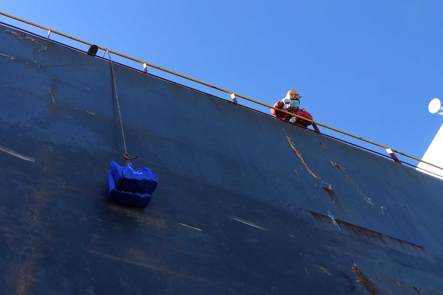 A man looks over the side of a ship as a bag of groceries is raised on a rope