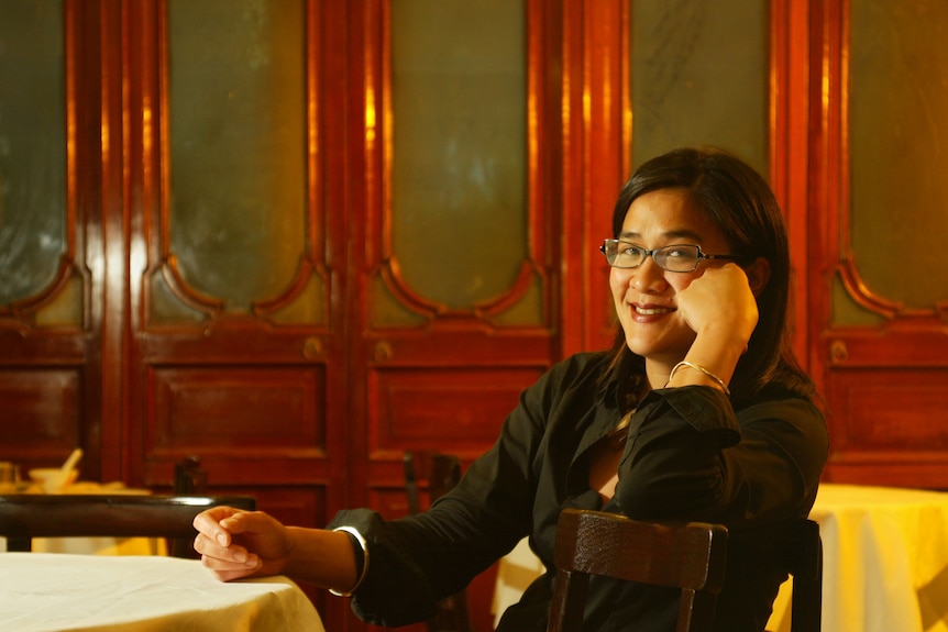 Kylie Kwong sits with hand on side of her face, sitting in a tea house restaurant with glass trim red doors behind her