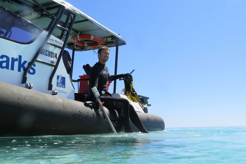 A man in a full wetsuit sits on the side of a boat.