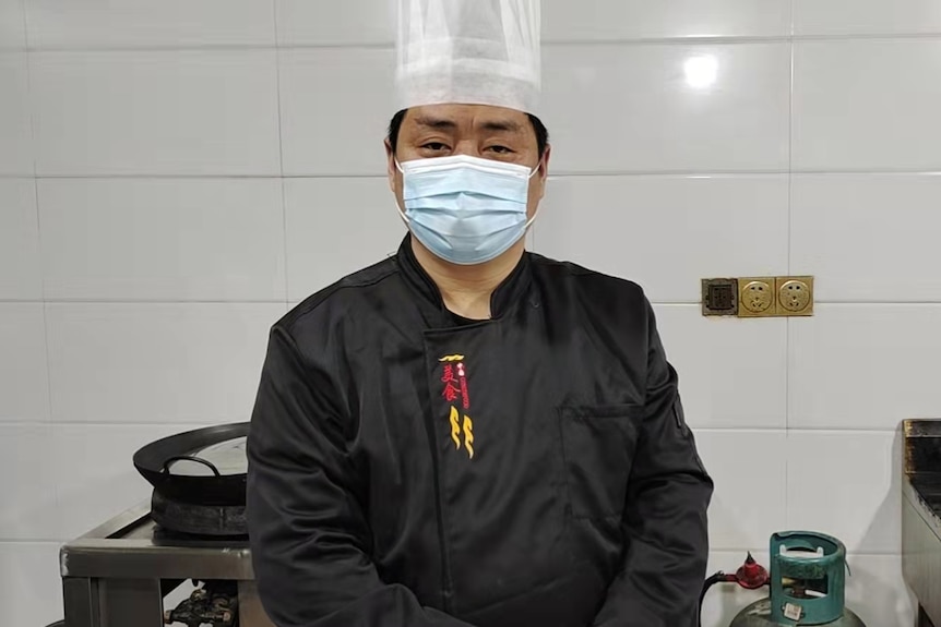 A man in a chefs hat wearing a mask look in a kitchen looks at the camera