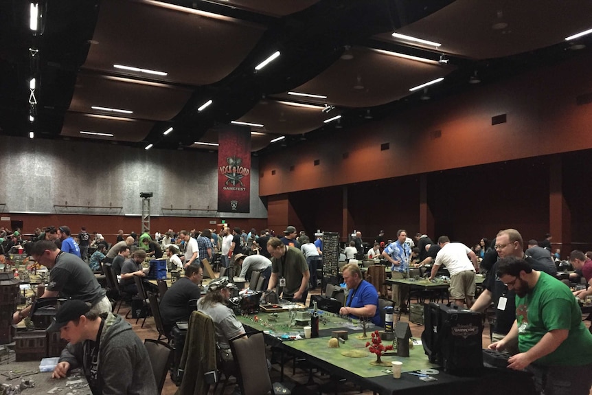 A room full of men sitting and standing at tables playing boardgames.