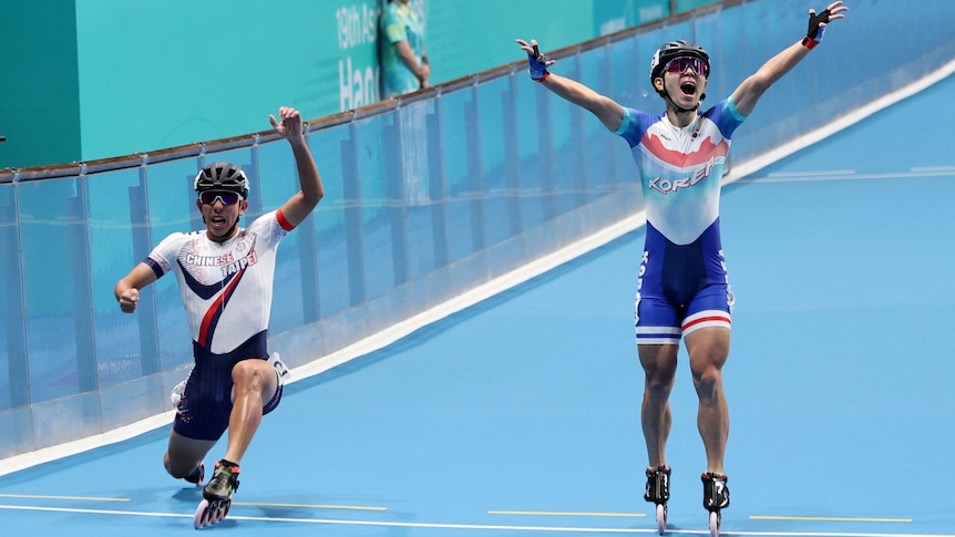 Two roller skaters celebrate as they cross a finish line. The one on the left is kneeling with his foot outstretched.