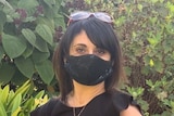 A woman with dark hair wearing a black shirt, black pencil skirt and black face mask stands in a well-manicured garden.