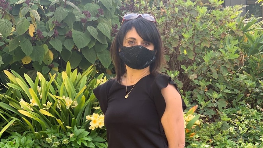 A woman with dark hair wearing a black shirt, black pencil skirt and black face mask stands in a well-manicured garden.