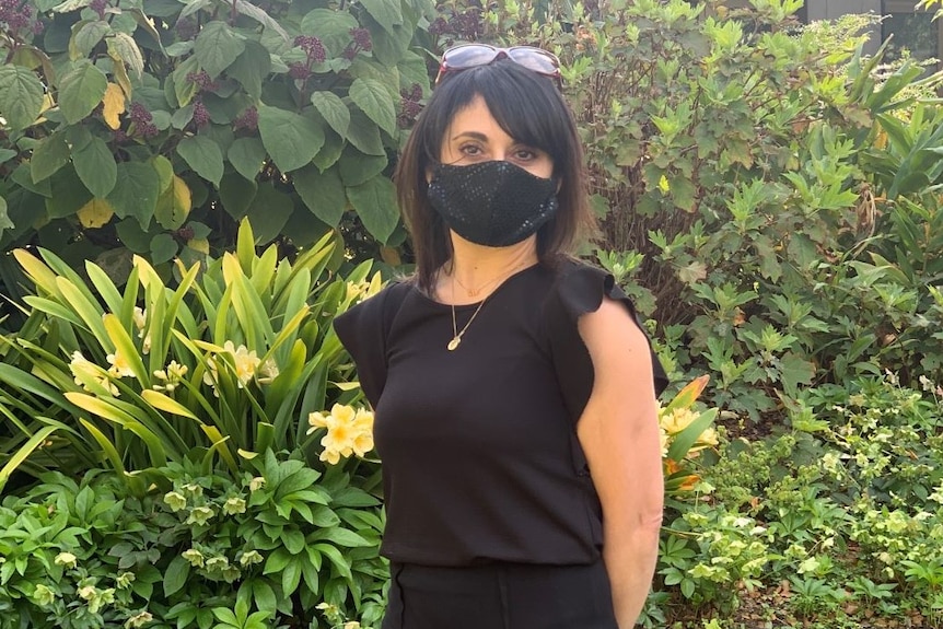 A woman with dark hair wearing a black shirt, black pencil skirt and black face mask stands in a well-manicured garden.