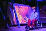 a man in a wheelchair sits in a barbecue shelter, illuminated by blue light and there's an image of a hand behind him 