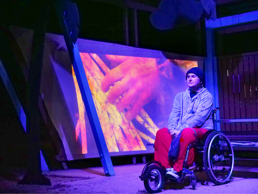 a man in a wheelchair sits in a barbecue shelter, illuminated by blue light and there's an image of a hand behind him 