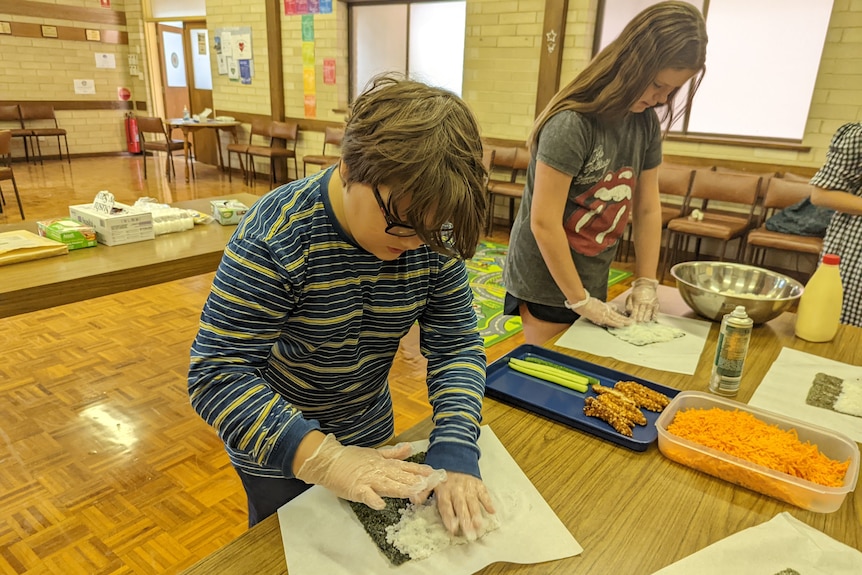 A young boy with brown hair, glasses and long-sleeved stripy top trying to put rice on seaweed to make sushi.