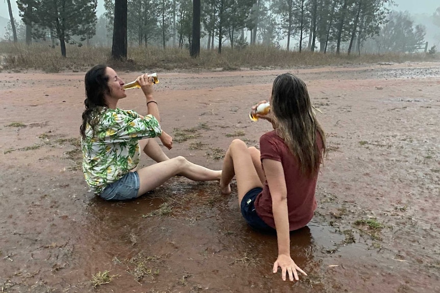Two women sit on a muddy road in the rain drinking with trees in the background and misty rain