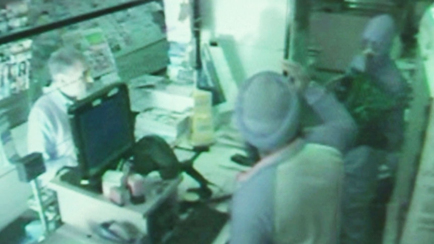 A man, wearing a bandana around his face, robs a post office worker.