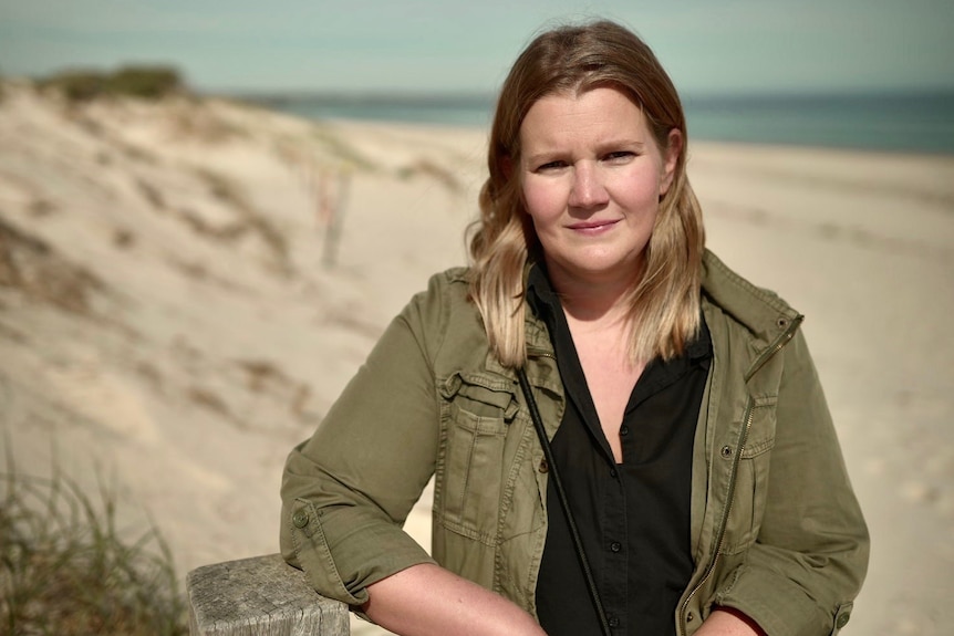 A woman wearing a black shirt under a green jacket stands on a beach.