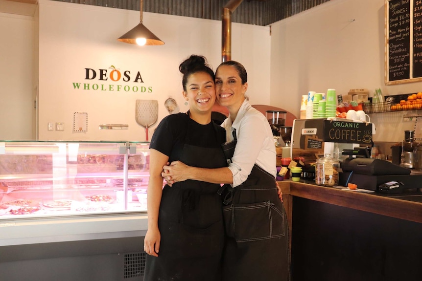 Two women in aprons hug in front of a coffee and deli counter inside a shop that has 'Deosa Wholefoods' written on the wall.