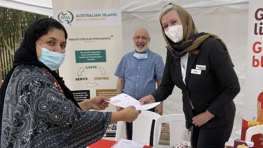 Two women wearing head scarves and a man stand at a blood donation table.
