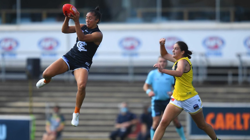 An AFLW footballer flies high to grab the ball in mid-air, while a defender chases after her.