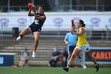 An AFLW footballer flies high to grab the ball in mid-air, while a defender chases after her.