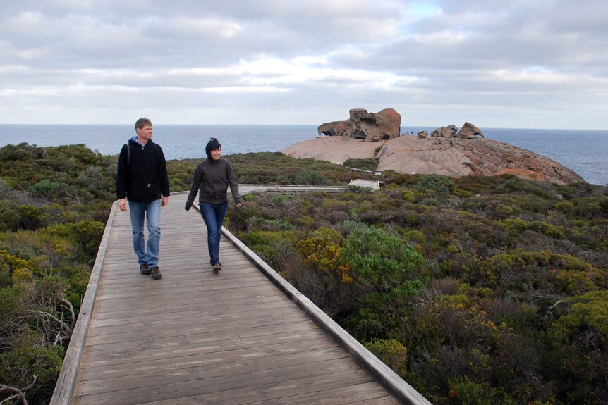 Two people walk on a boardwalk above green shrubbery, with large rocks in the background.