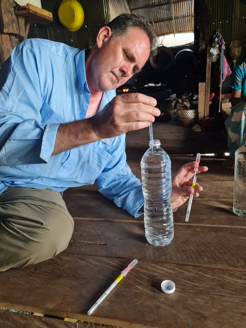 A serious middle-aged man in a light blue shirt, khaki pans, sits on a wooden board or desk, puts syringe into a plastic bottle.