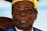 Close-up of tired-looking Mugabe in gown and mortarboard with military officer saluting behind him.