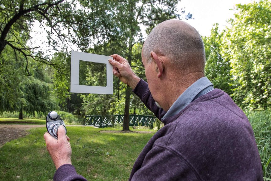 A man holds up a cardboard frame and a light meter in a park of trees and green grass