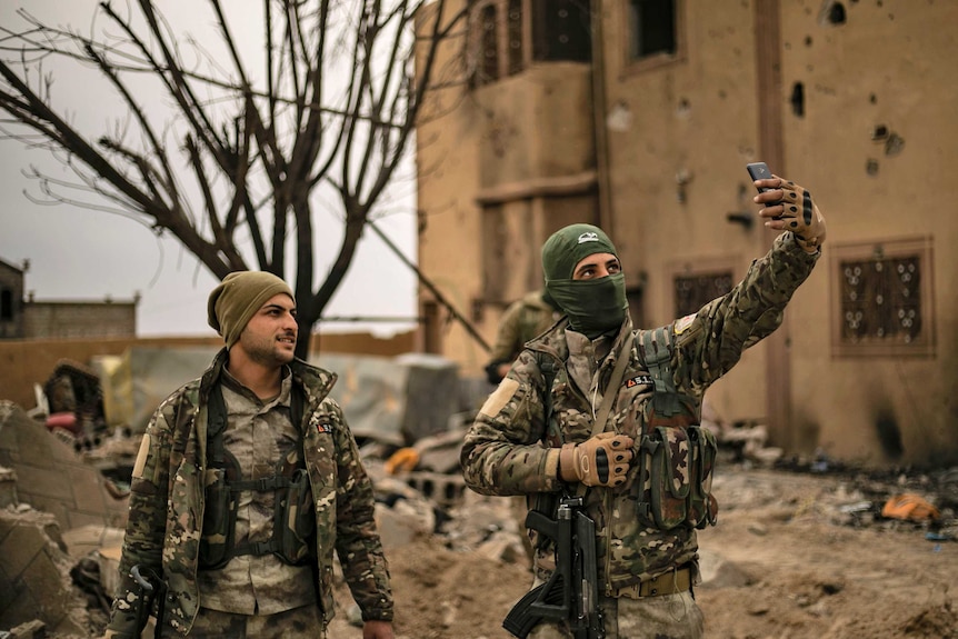 Soldiers in Syria in uniform smile for an iphone amidst rubble.