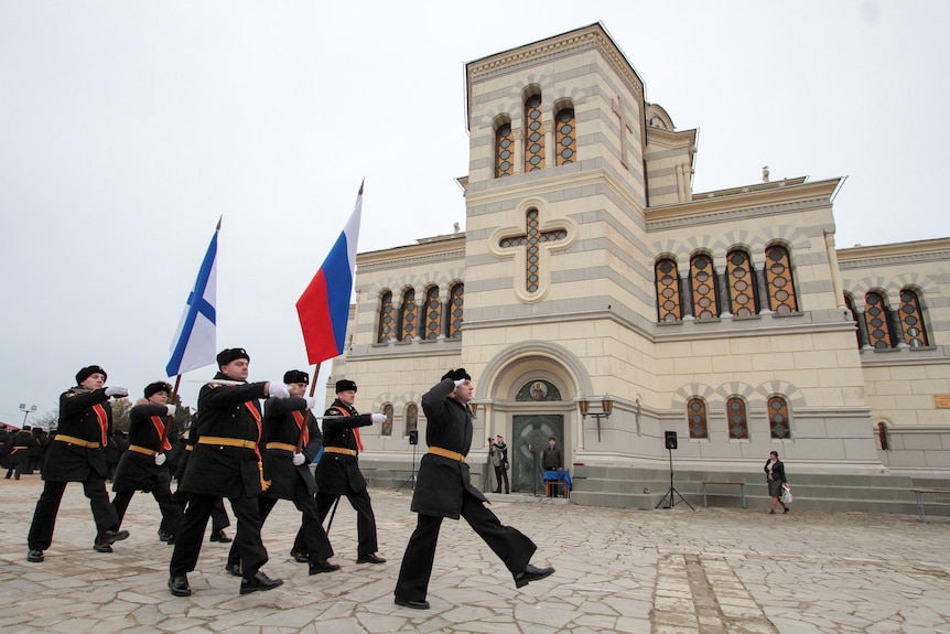 A group of black-clad sailors march past a white stone cathedral holding Russian flags.