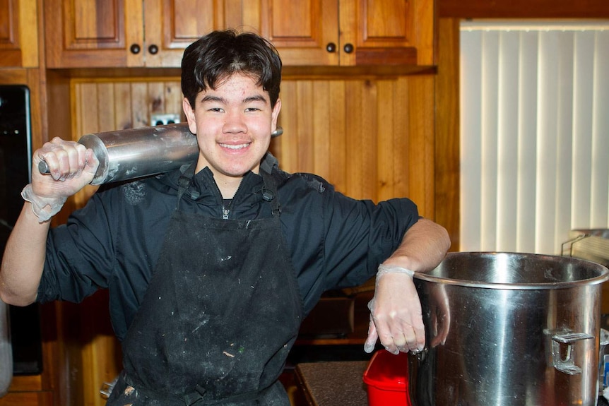 Smiling teenager with industrial rolling pin over his shoulder leaning on a huge mixing bowl