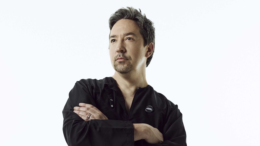 A profile shot of a man with black hair wearing a black shirt with his arms folded.