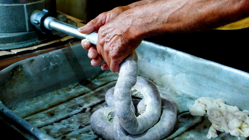 Hands with prominent veins hold the end of a tube, which releases a white rope of uncured chorizo that pools in a metal tin.