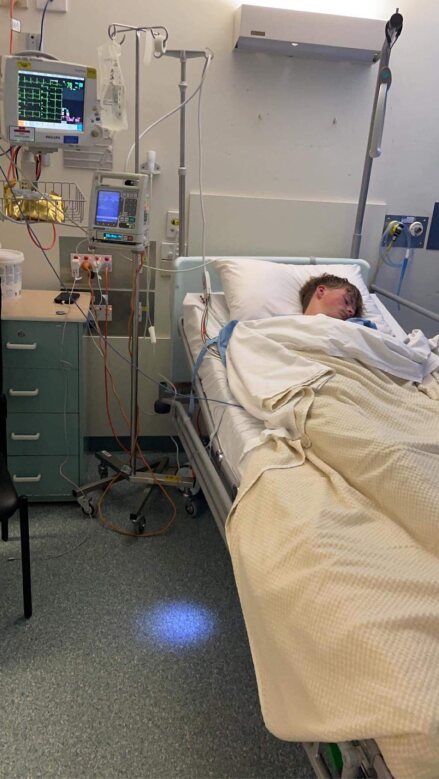 Malik lies unconscious in a hospital bed connected to oxygen and other machines.