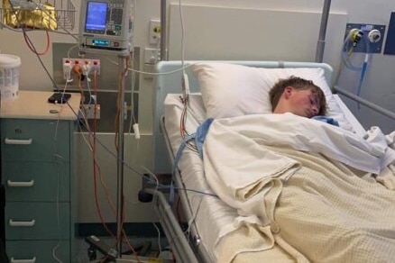 Malik lies unconscious in a hospital bed connected to oxygen and other machines.