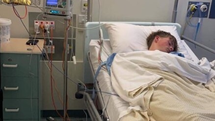 Malik lies unconscious in a hospital bed connected to oxygen and other machines.