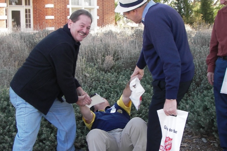 A smiling man leans over to help a another man, who is joking and pretending to be tired, stand up from the ground.