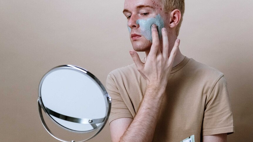 A man applies a mud mask to his face while looking in a mirror
