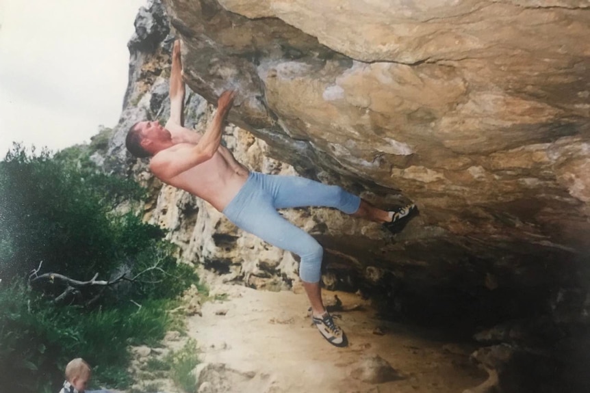 A shirtless man scales an overhanging rock, not far from the ground.
