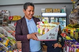 A man stands in a grocery store smiling, holding a box of baby toys.