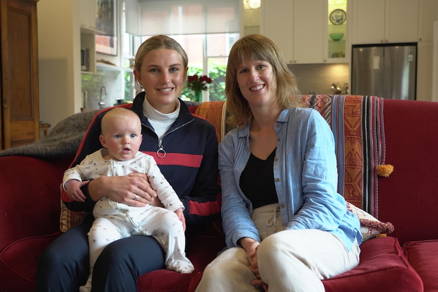Two young women sit on a red couch, one of them holds a six-month-old baby