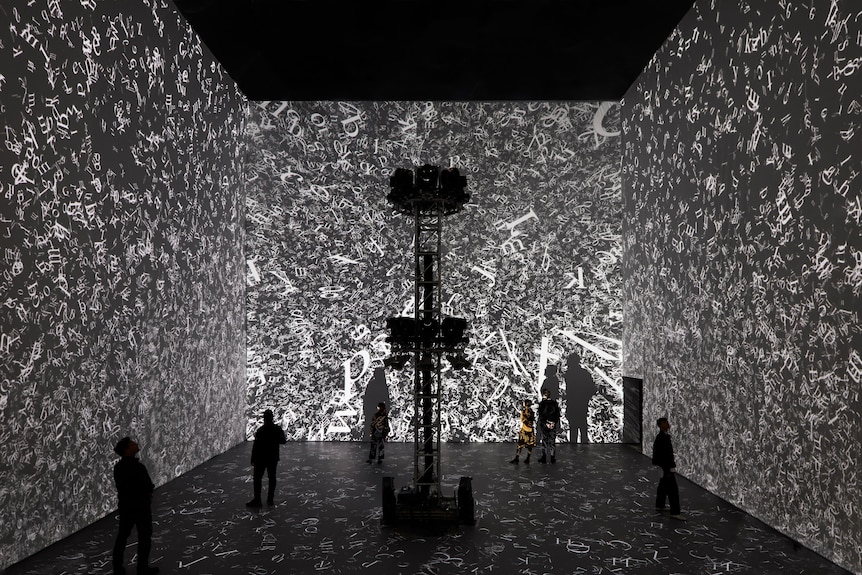 A group of people wander through a large gallery space with wall-to-wall projections of black and white text.