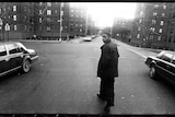 A black and white image of Nas in the middle of a street looking back at the camera, two cars on either side