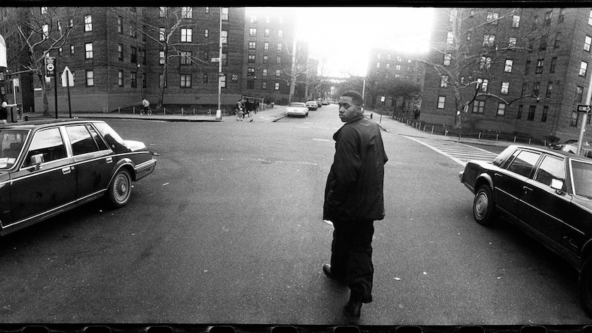 A black and white image of Nas in the middle of a street looking back at the camera, two cars on either side