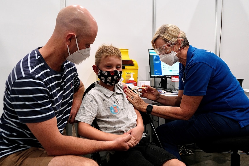 A young boy with blonde hair receives an injection to his arm while his father holds his hand.