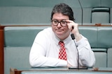 A dark-haired, bespectacled man wearing a crisp white shirt and a red tie sitting in federal Parliament.