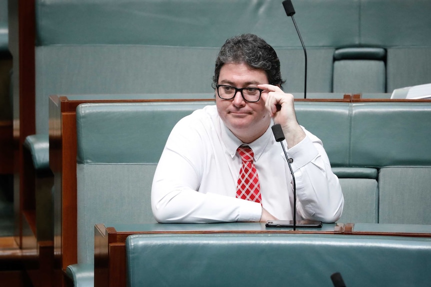 A dark-haired man sitting, leaning forward in the federal House of Representatives, adjusting glasses.